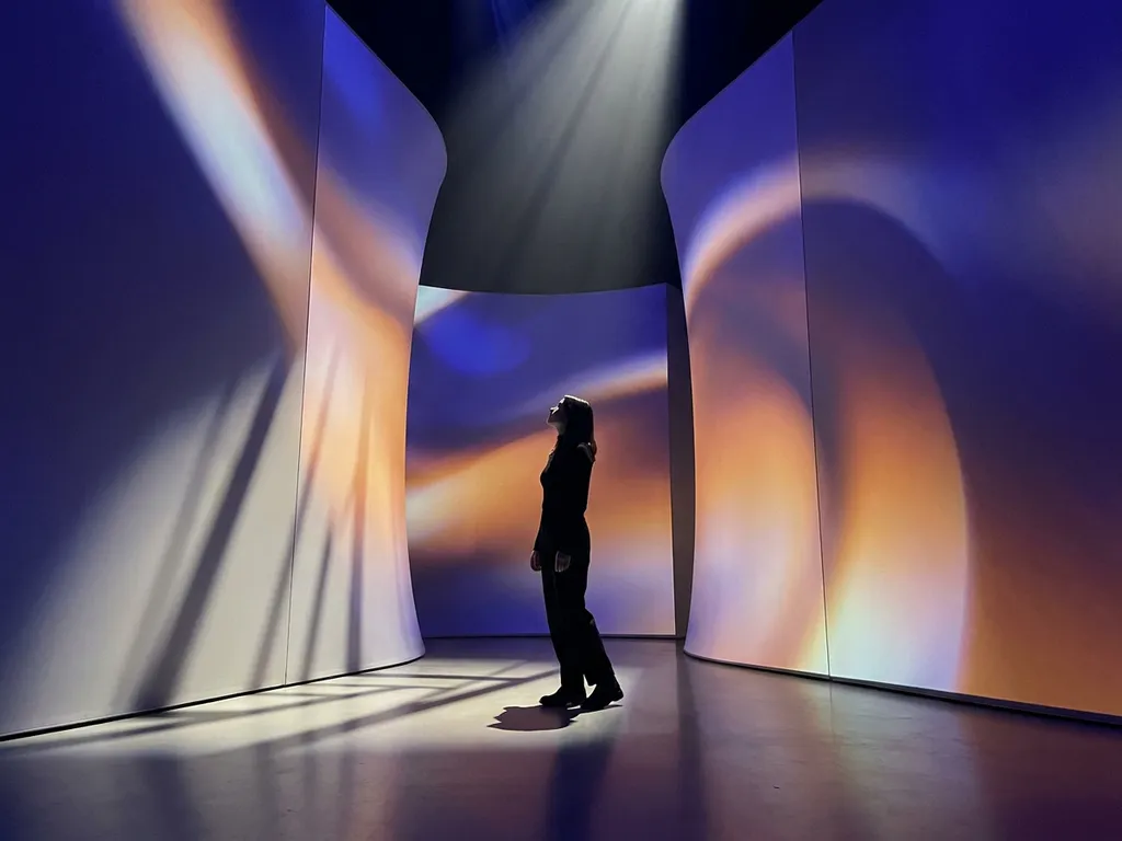 Visitor standing inside an immersive light installation, curved walls bathed in deep indigo and warm amber projected light, cinematic low-angle perspective.