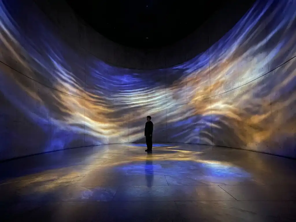 Solitary visitor dwarfed by sweeping indigo and amber light projections flowing across curved walls of an immersive art installation.