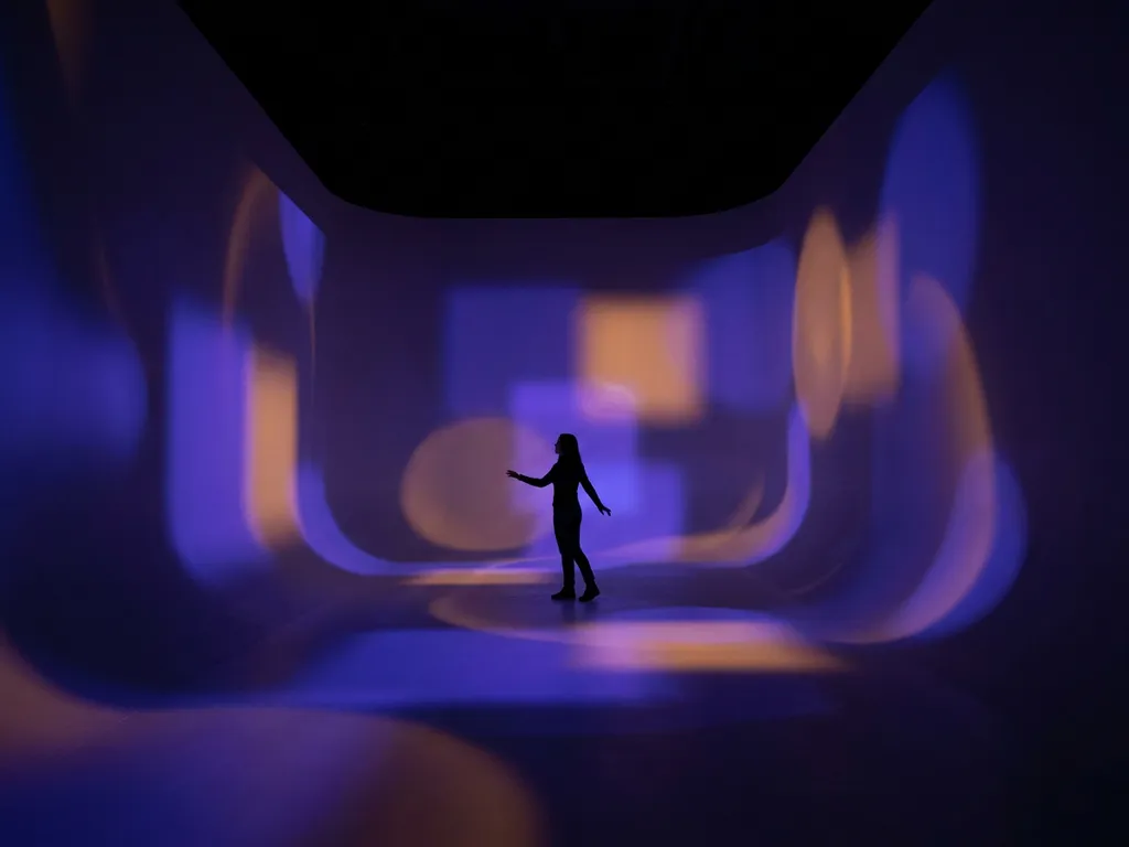 Lone visitor standing in an immersive darkened room with indigo and amber light projections washing across curved white walls.