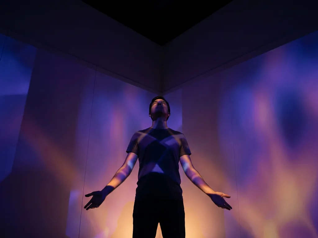 Person standing in immersive projection room, hands outstretched as indigo and amber geometric light patterns wash over skin and walls.