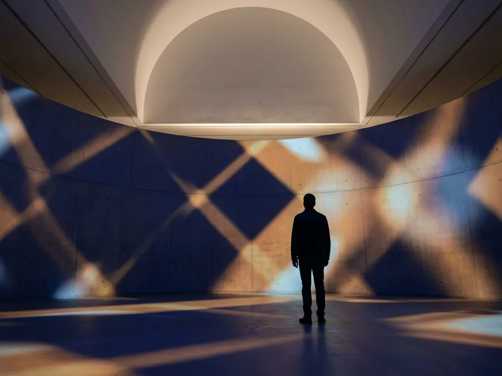 Person standing in an immersive projection room with navy blue and amber light wrapping the walls and floor in minimal geometric forms.