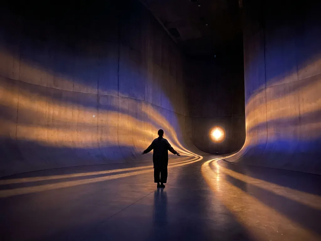 Lone visitor standing in a darkened immersive art installation with amber and indigo projected light washing over curved walls.