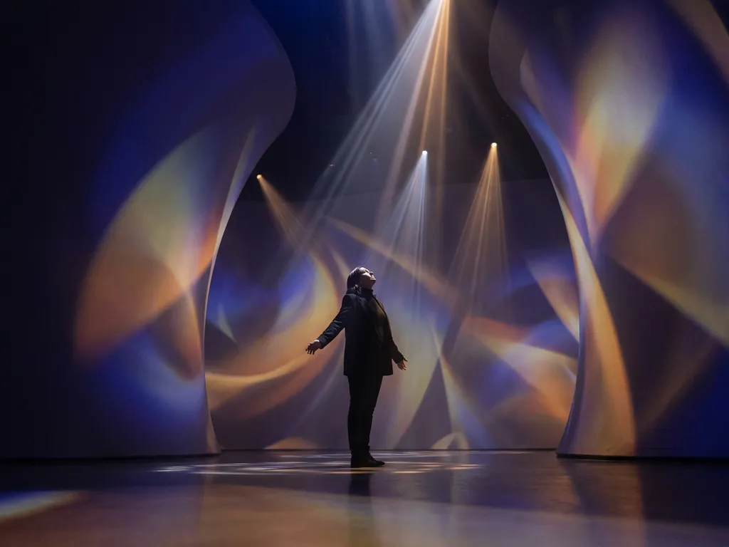 Visitor standing in an immersive dark room with indigo and amber projected light washing across curved walls.