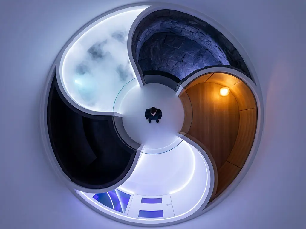 Person at center of circular room with five glowing sensory zones radiating outward in indigo, white, and amber tones, overhead view.