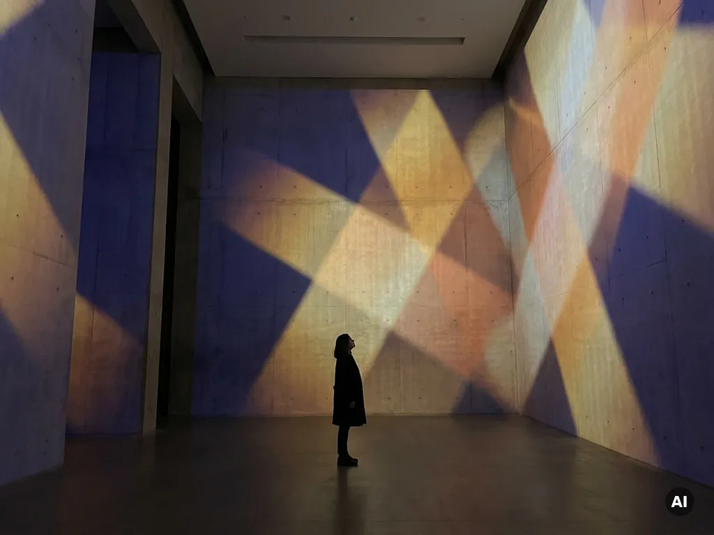Lone visitor standing in a darkened gallery surrounded by floor-to-ceiling navy and amber projected geometric light patterns on concrete walls.